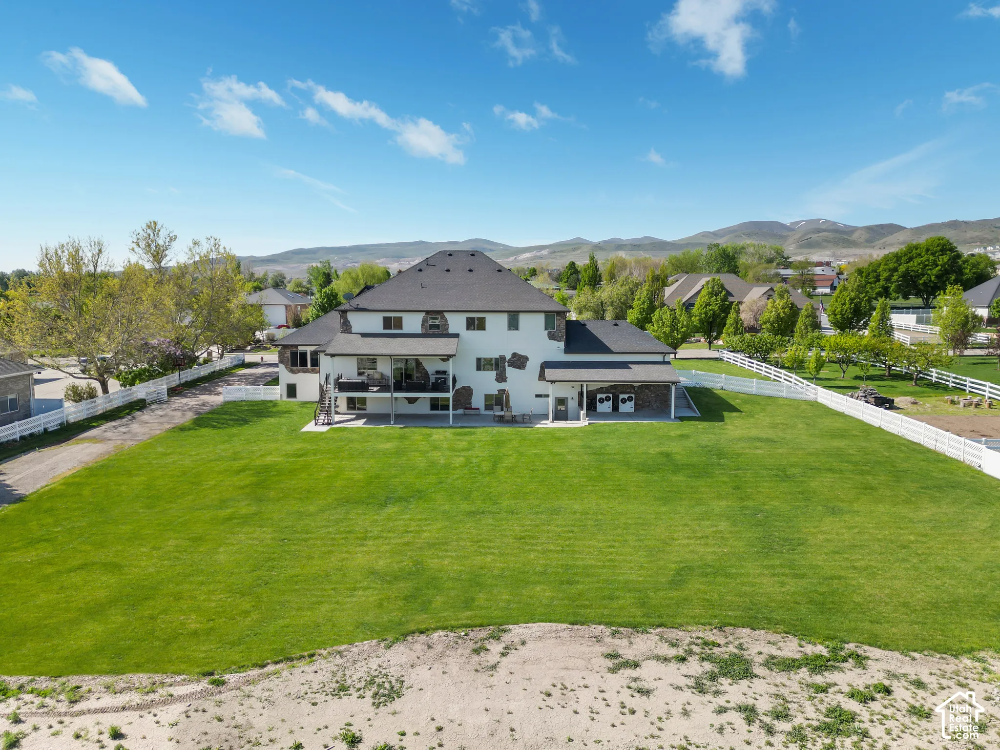 Expansive backyard featuring a manicured lawn, white vinyl fencing, covered patio, stone veneer accents, and mountain backdrop