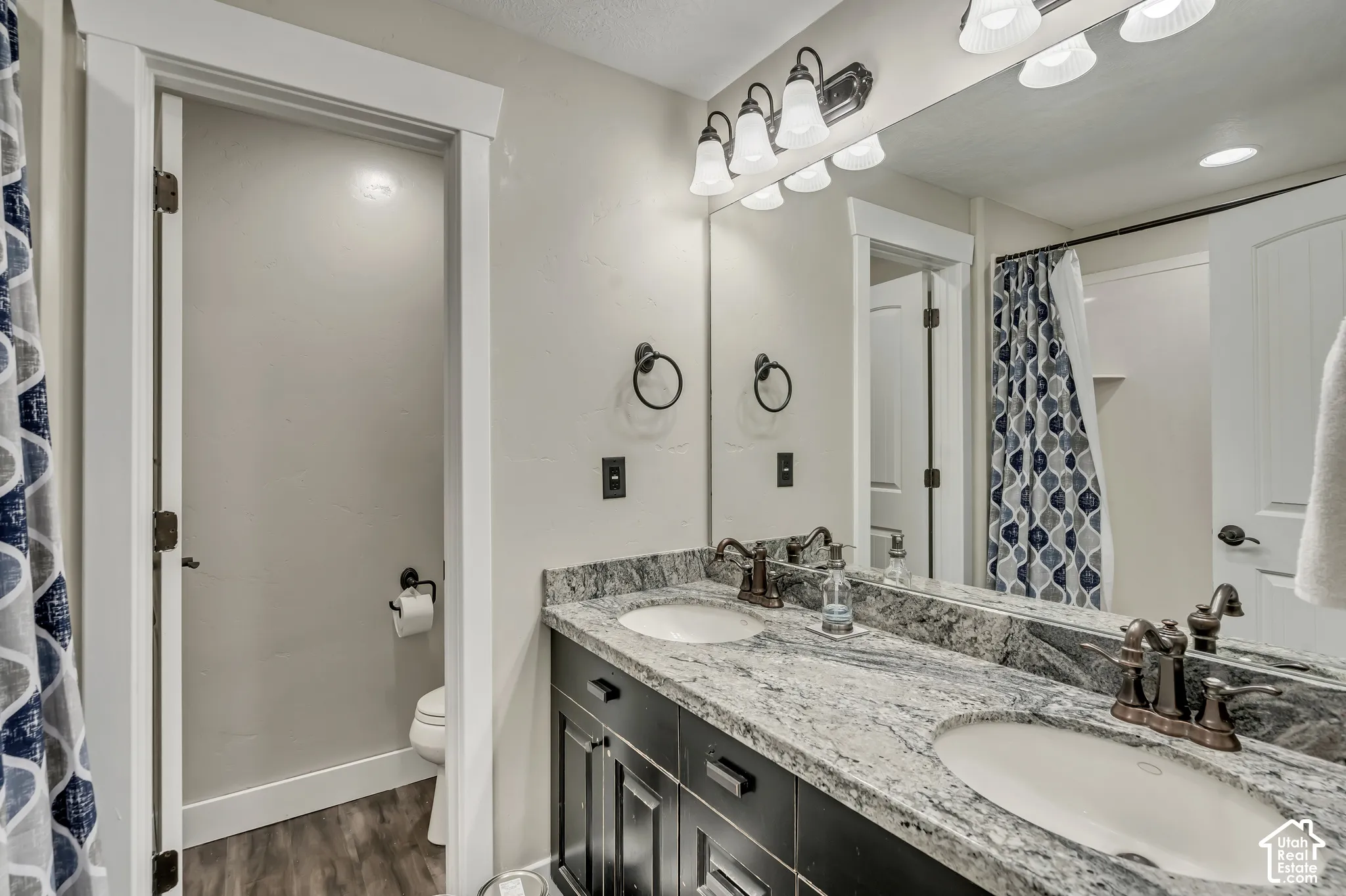 Dual-sink vanity with stone countertop, dark cabinetry, oil-rubbed bronze fixtures, full-width vanity mirror, and three-light sconce