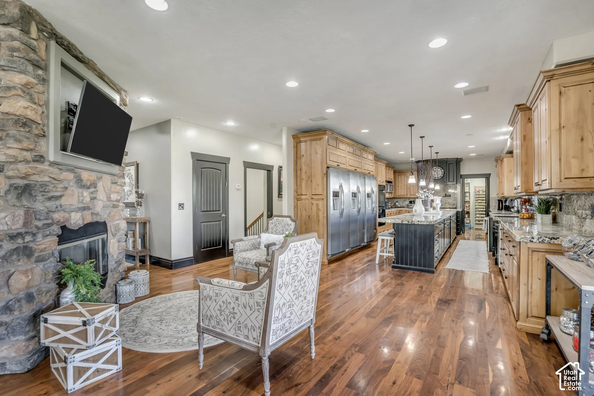 Floor-to-ceiling natural stone fireplace with a recessed television nook, wood-finish flooring, custom wood cabinetry, stainless steel professional-grade refrigerator, and expansive central island with stone countertop