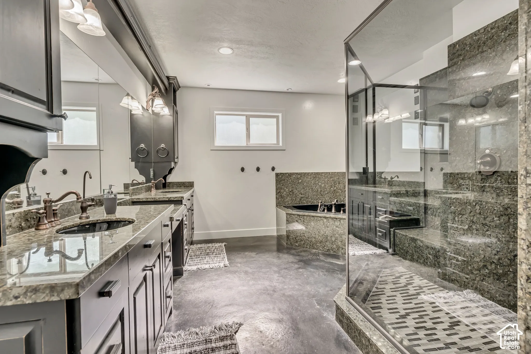 Bathroom featuring dual-sink vanity with stone countertops, expansive walk-in glass shower with integrated bench seating, and a deep soaking tub surrounded by stone tile