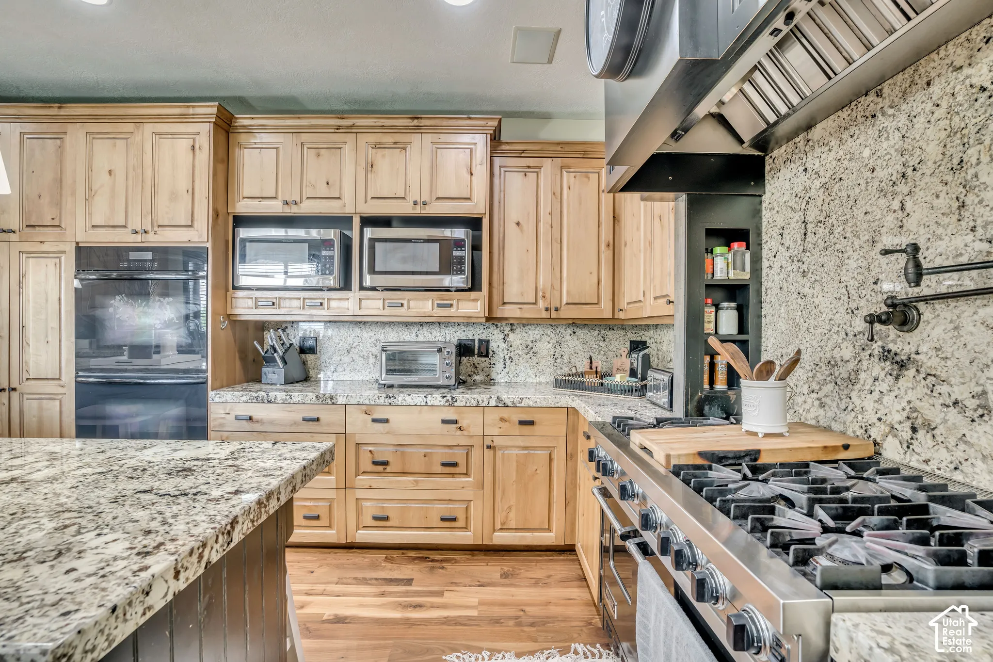 Kitchen featuring natural wood cabinetry, stone-surface countertops, professional-grade stainless steel gas range with a wall-mounted pot filler, built-in stainless steel double oven, and wood-finish flooring