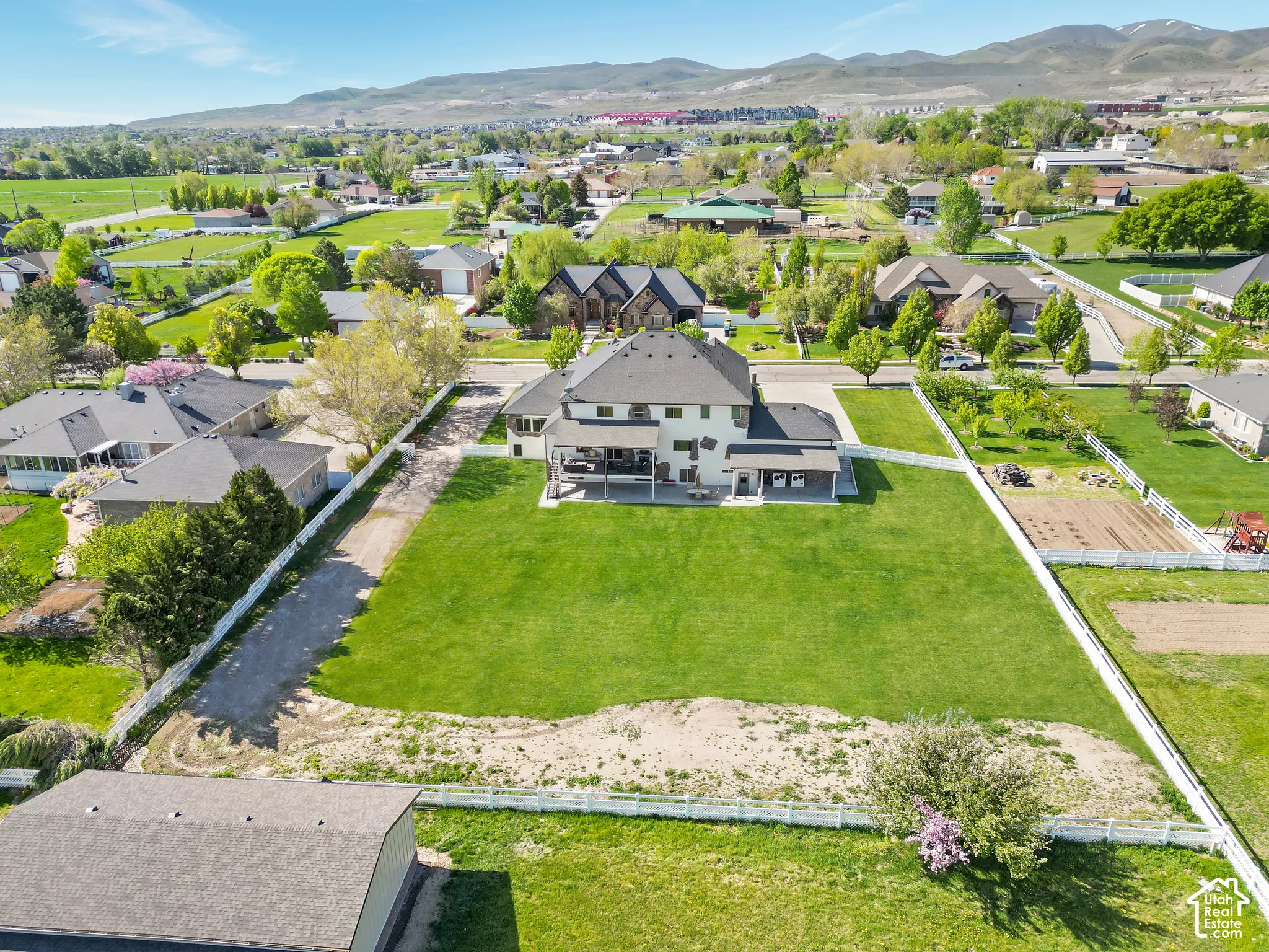 Aerial perspective of a multi-story residence featuring a covered patio, extensive open lawn, perimeter white vinyl fencing, and mountain backdrop