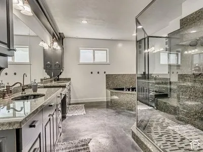 Bathroom featuring dark-toned cabinetry, stone-finish countertops with dual undermount sinks, a glass-enclosed shower, and a built-in soaking tub with stone surround