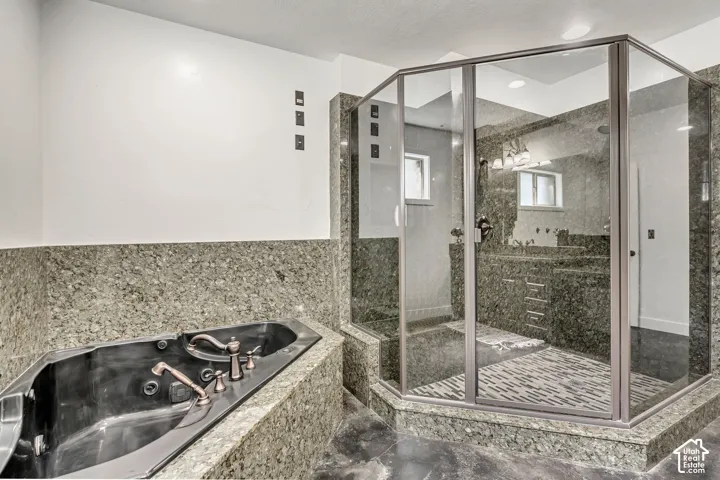Built-in jetted tub with metallic fixtures, full-surround stone tiling, corner glass-enclosed shower, stone flooring, and recessed ceiling lighting