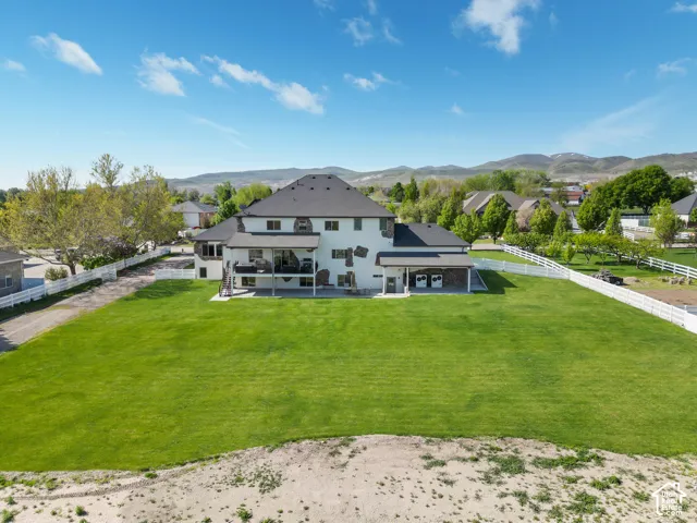 Expansive backyard featuring a manicured lawn, white vinyl fencing, covered patio, stone veneer accents, and mountain backdrop