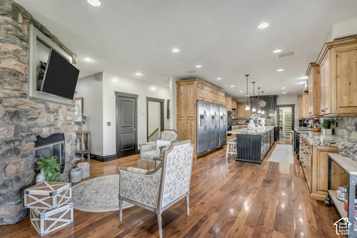 Floor-to-ceiling natural stone fireplace with a recessed television nook, wood-finish flooring, custom wood cabinetry, stainless steel professional-grade refrigerator, and expansive central island with stone countertop