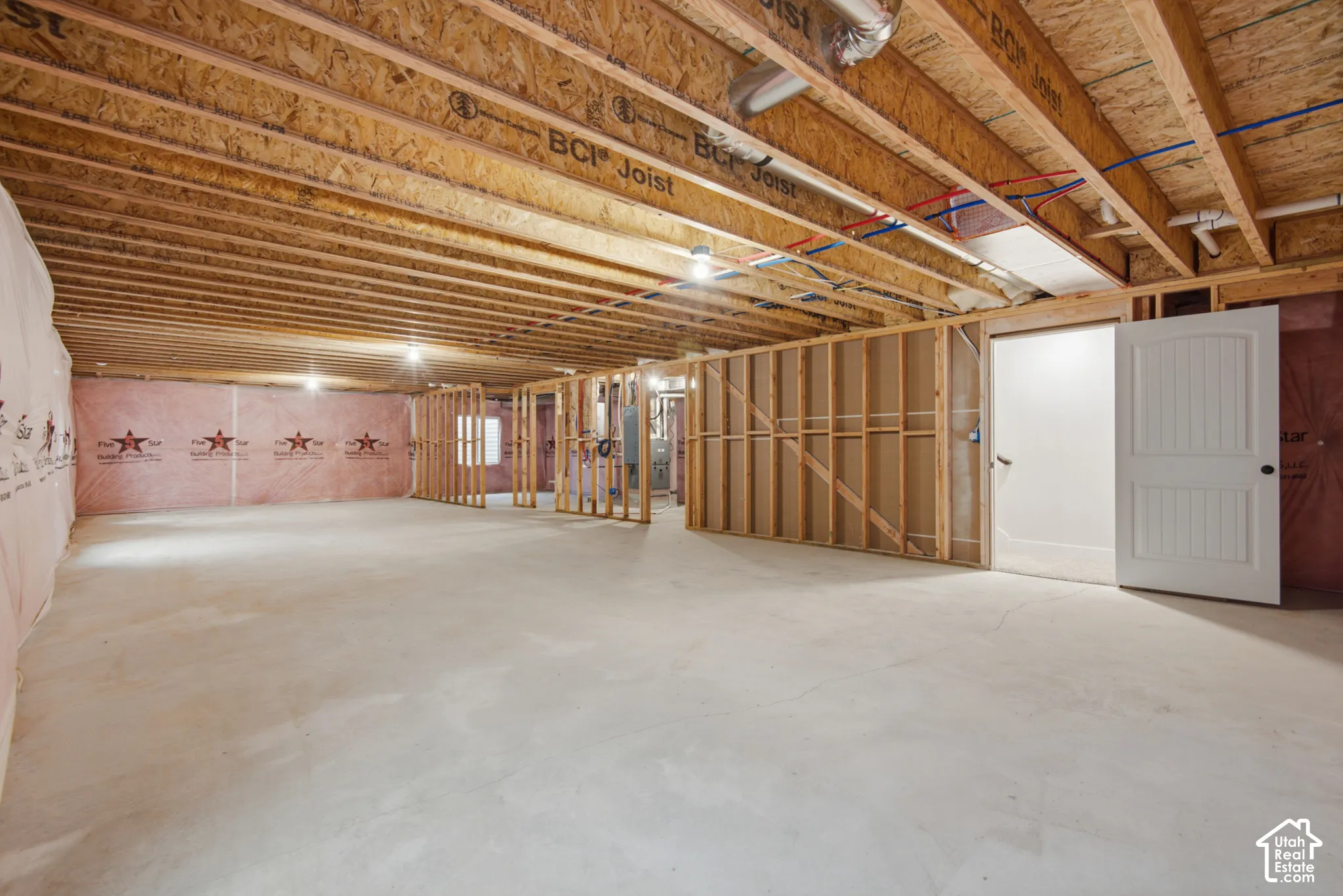 Unfinished basement space featuring exposed engineered joists, concrete flooring, pink fiberglass insulation, and wood-framed partition walls