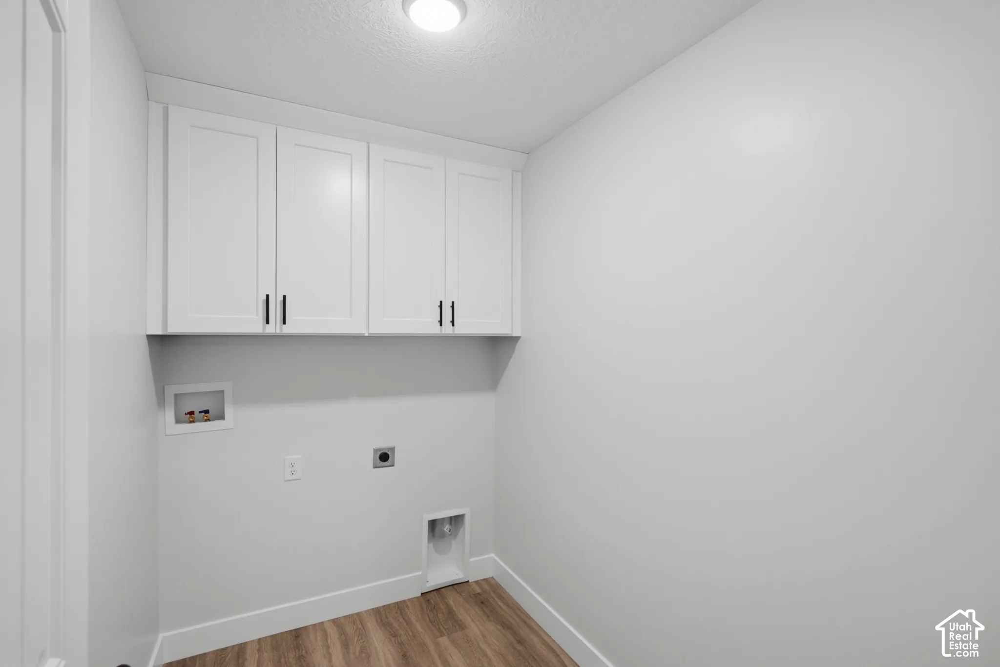 Utility room featuring white shaker-style wall cabinets with matte black hardware, recessed washer and dryer hookups, and wood-finish flooring