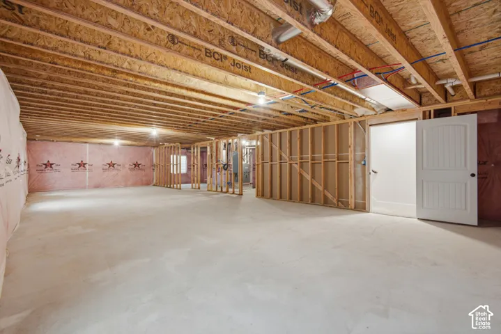 Unfinished basement space featuring exposed engineered joists, concrete flooring, pink fiberglass insulation, and wood-framed partition walls