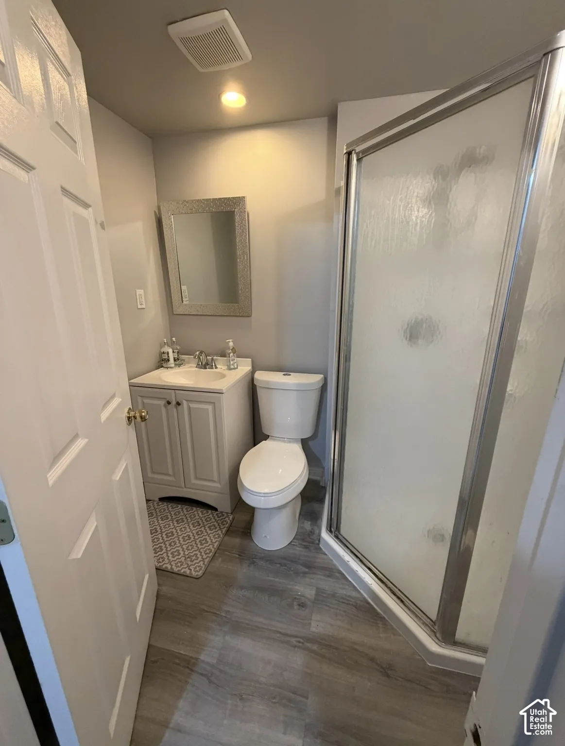 Bathroom featuring wood-finish flooring, a corner glass-enclosed shower, a white vanity with a composite countertop, and a wall-mounted rectangular mirror