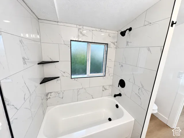 Bathtub surround featuring large-format white stone-finish tile, matte black fixtures, built-in corner shelving, and an obscured-glass window