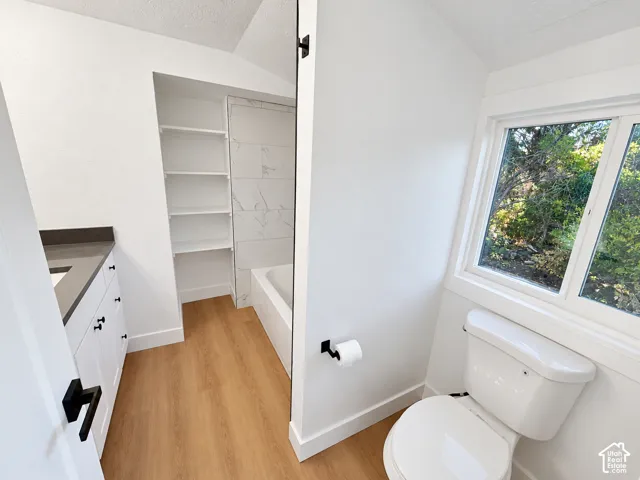White vanity with dark countertop, wood-finish flooring, built-in shelving, bathtub with tile surround, and matte black hardware