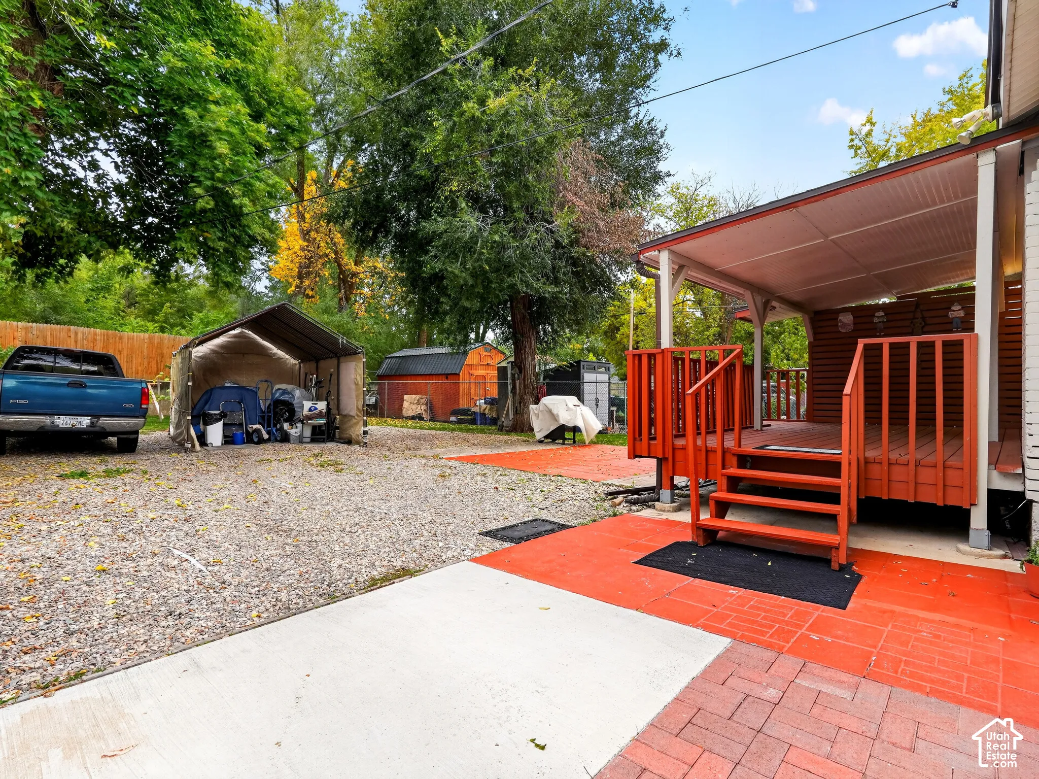 Covered deck with wooden railing and stairs, brick-paver patio, gravel parking area, and mature trees
