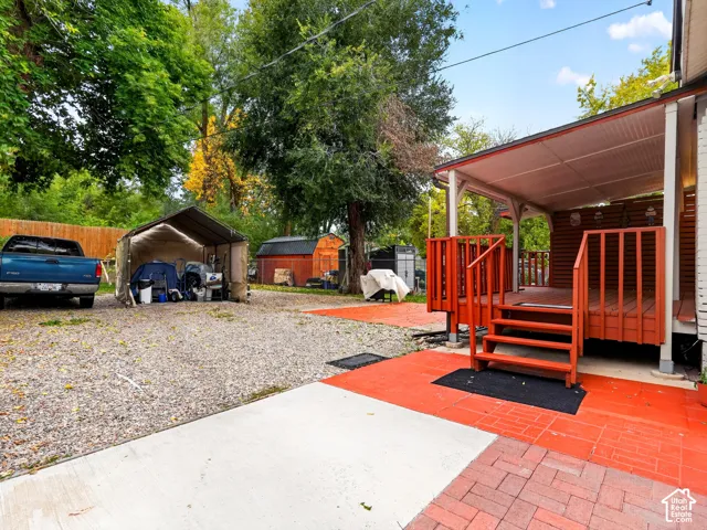 Covered deck with wooden railing and stairs, brick-paver patio, gravel parking area, and mature trees