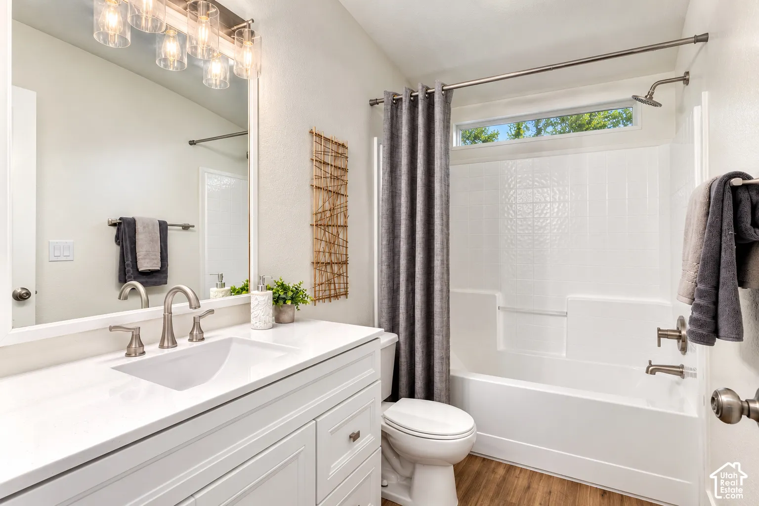 Bathroom featuring a white vanity with a rectangular undermount sink, quartz countertop, multi-light fixture, wood-finish flooring, and a tub-shower combination with a transom window