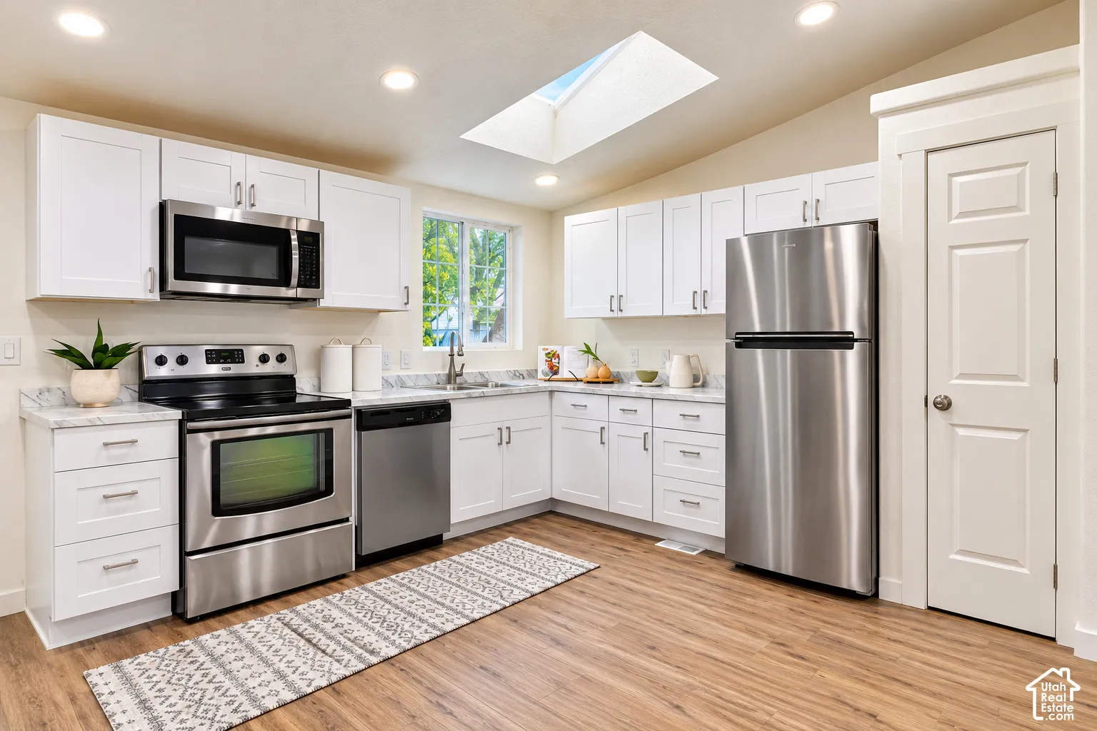 Kitchen featuring white shaker-style cabinetry, stainless steel appliances, stone-patterned countertops, a skylight, and wood-finish flooring