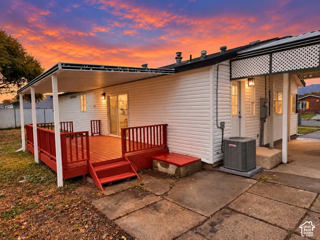Exterior featuring white horizontal siding, a covered wooden deck with railing, sliding glass doors, a concrete patio, and a carport with decorative lattice trim