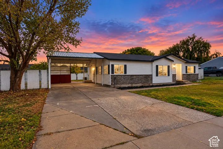 Single-story exterior featuring white horizontal siding, stone veneer accents, a concrete driveway, an attached carport, and a chain-link fence