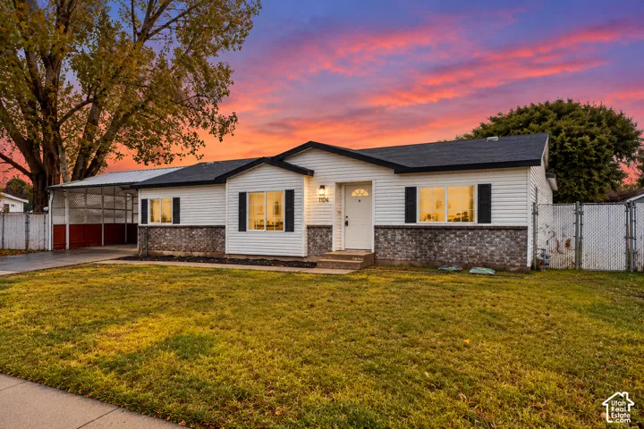 Single-story residence featuring white horizontal siding, a stone veneer base, a covered carport, and perimeter chain-link fencing