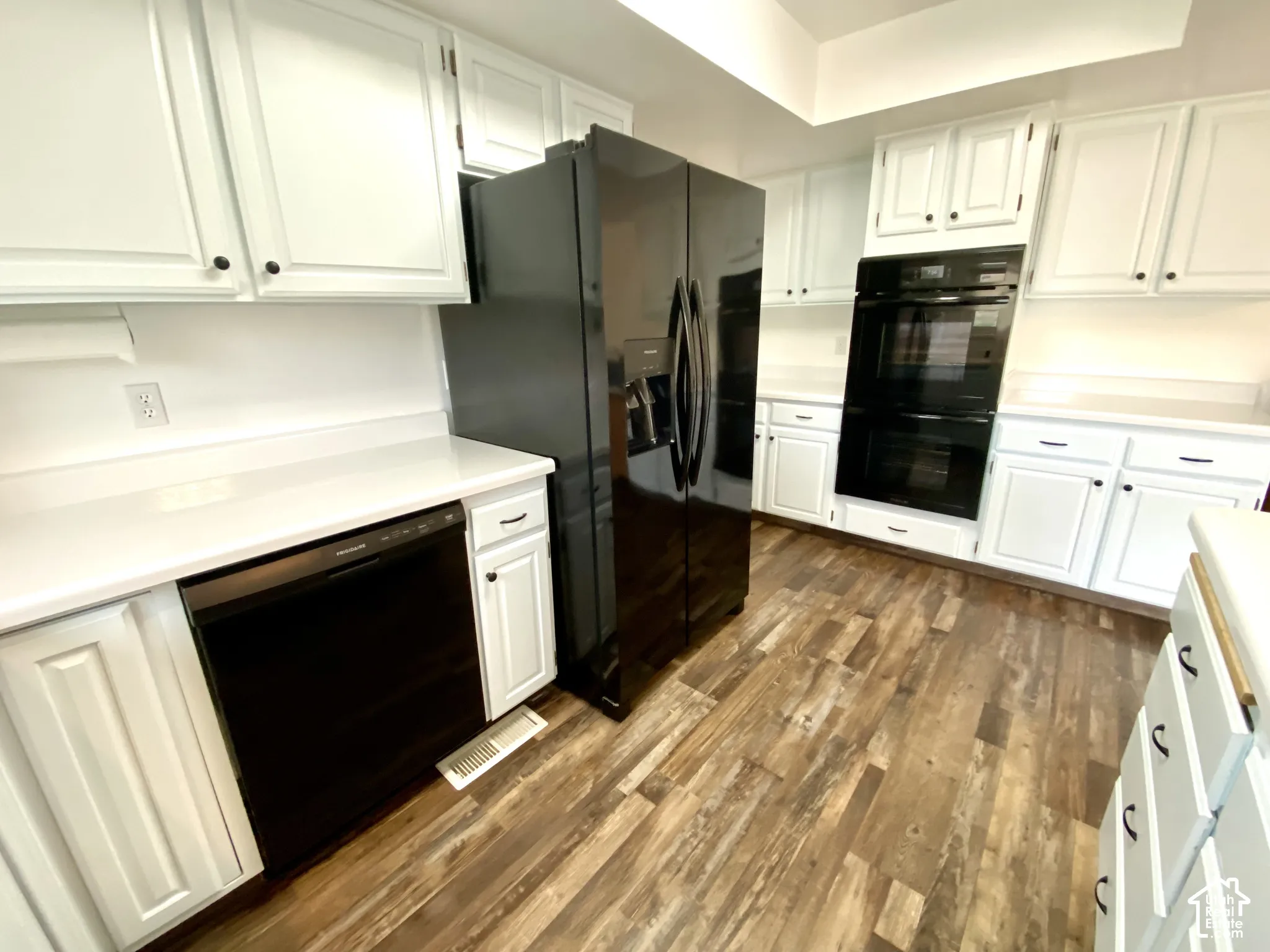 Kitchen featuring white shaker-style cabinetry, solid-surface countertops, wood-finish flooring, and black built-in appliances including a side-by-side refrigerator, dishwasher, and double wall oven