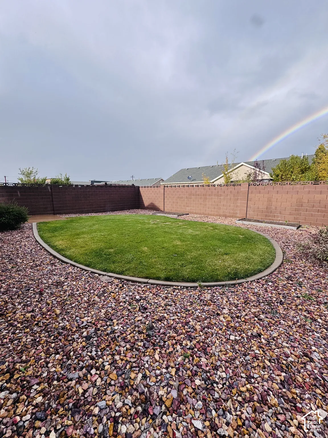 Backyard featuring a central oval lawn, concrete curbing, decorative rock ground cover, perimeter masonry wall, and built-in raised garden beds