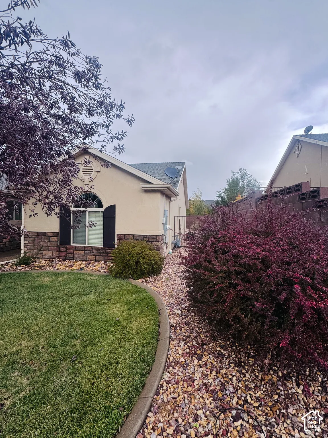 Exterior elevation featuring stucco siding, stone veneer accents, an arched window with decorative shutters, and landscaped grounds with gravel beds and a manicured lawn