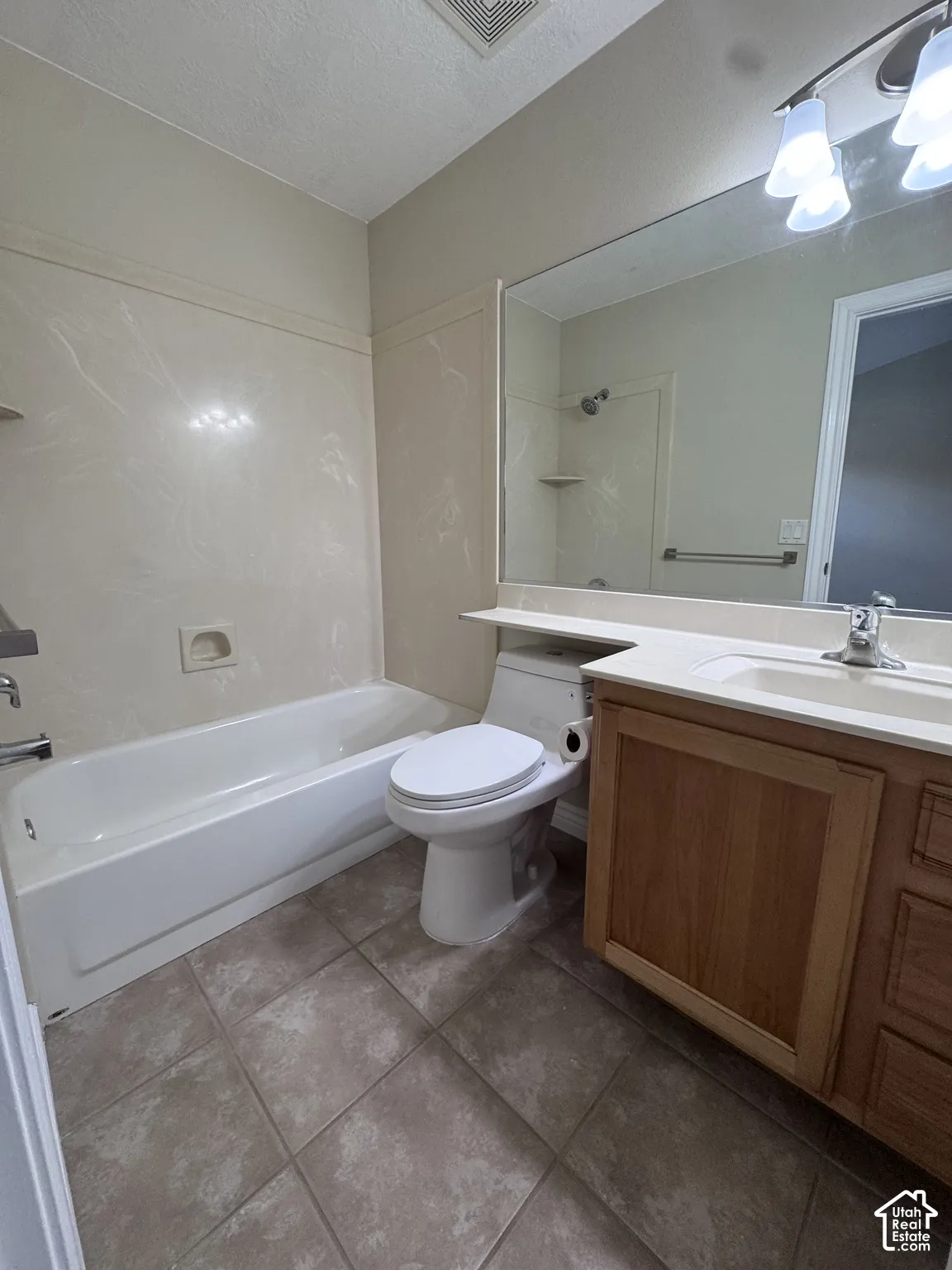 Bathroom featuring a wood-finish vanity with a light-toned countertop, a full-width wall mirror, a ceramic tile floor, and a combination tub and shower enclosure with surround paneling
