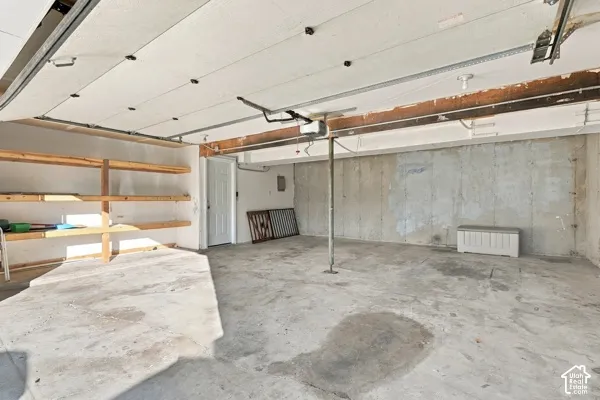 Enclosed garage featuring concrete flooring, exposed poured concrete walls, built-in wooden shelving, and a motorized garage door opener