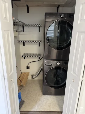 Laundry closet featuring stacked high-capacity front-load appliances, tiled flooring, and multiple wall-mounted shelving units