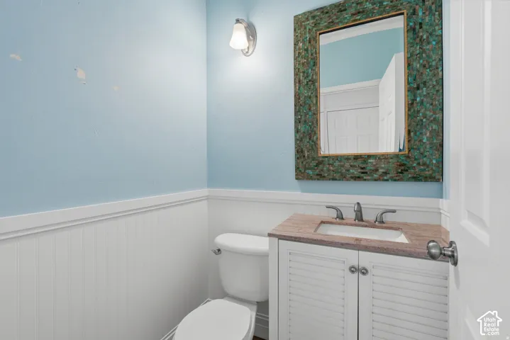 Vanity with a neutral-tone countertop and inset sink, shutter-style cabinet doors, wall-mounted light fixture, and decorative framed mirror featuring mosaic-style detailing, accented by white wainscoting