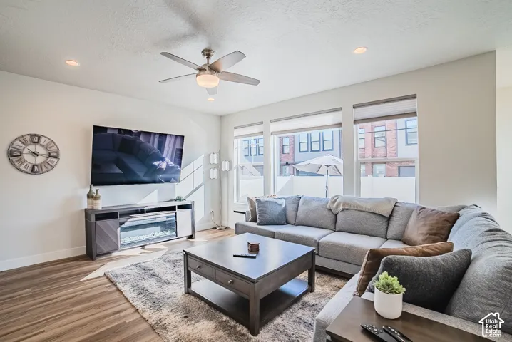 Living space featuring wood-finish flooring, recessed lighting, a brushed-metal ceiling fan, and large windows with blinds