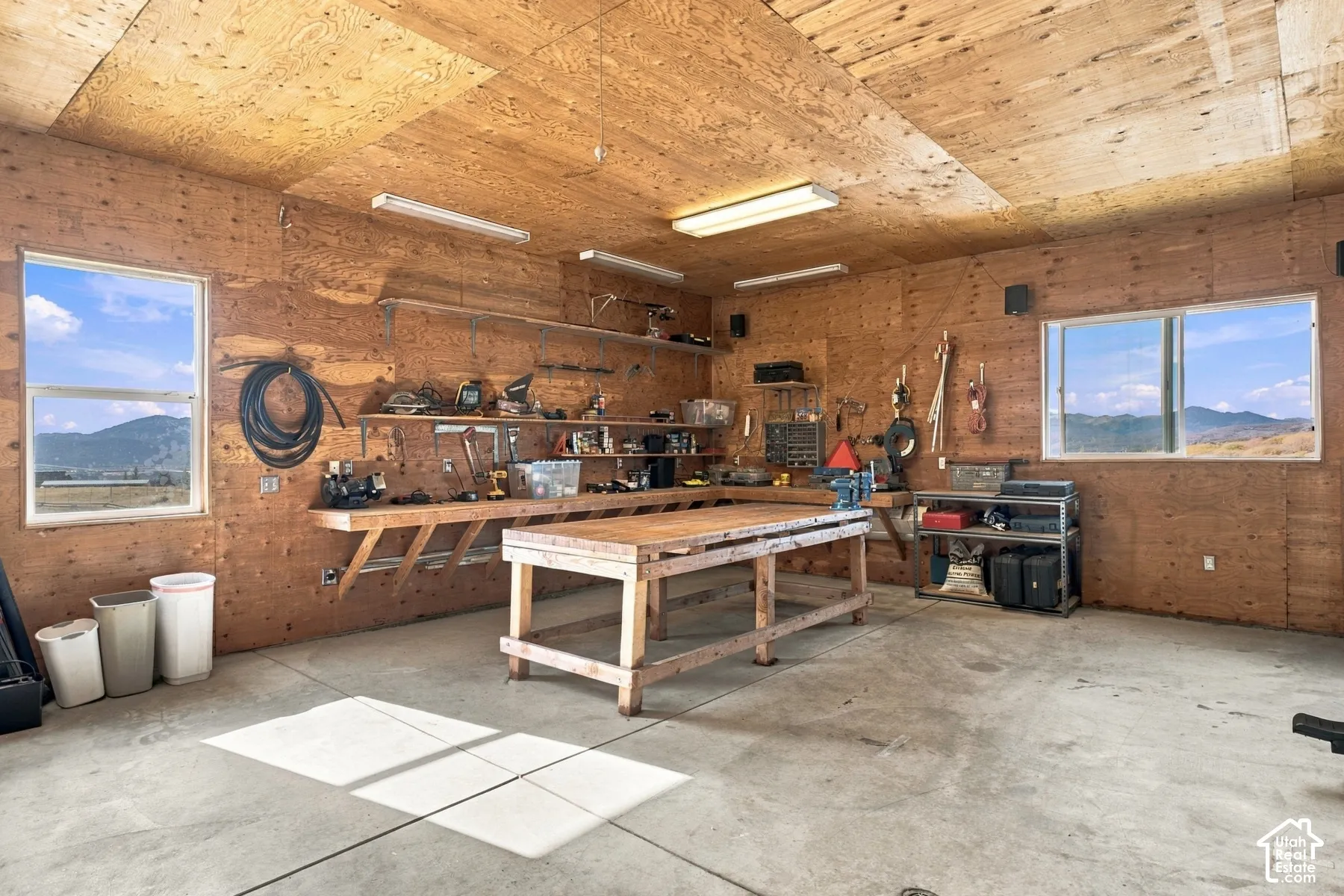 Workshop featuring wood-paneled walls, built-in wooden workbench, concrete flooring, fluorescent lighting, and dual windows