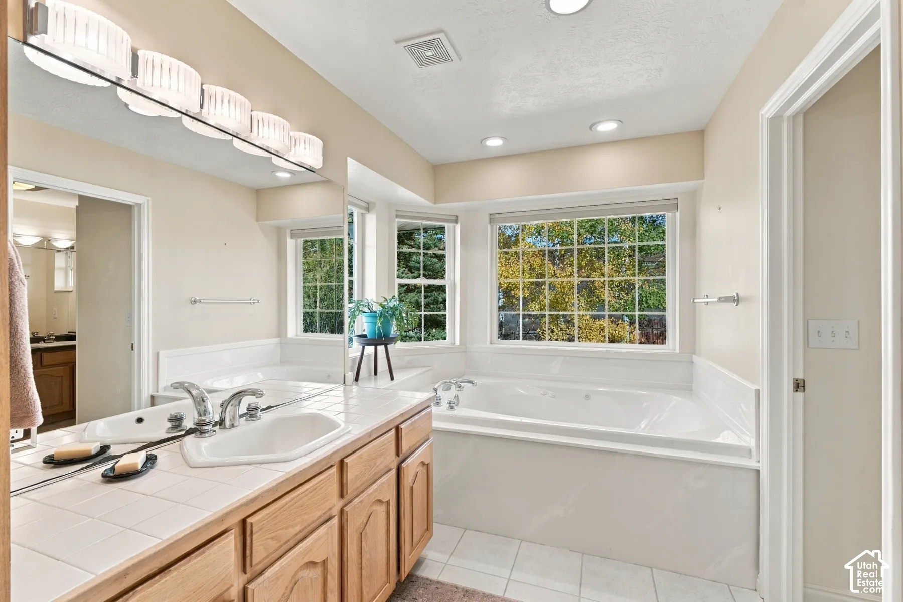 Bathroom featuring a large jetted soaking tub, tiled vanity countertop with an integrated basin, wood cabinetry, multi-bulb vanity lighting, and multiple windows