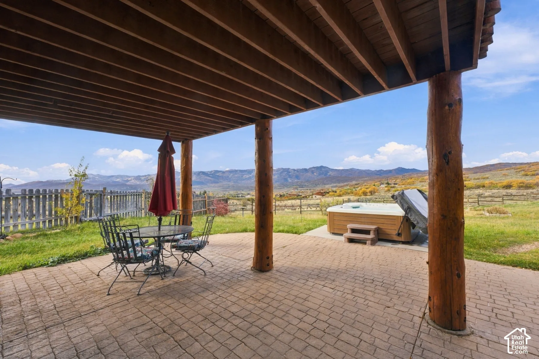 Covered patio with rustic log columns and overhead timber beams, featuring stamped concrete hardscaping, an integrated spa, and mountain scenery