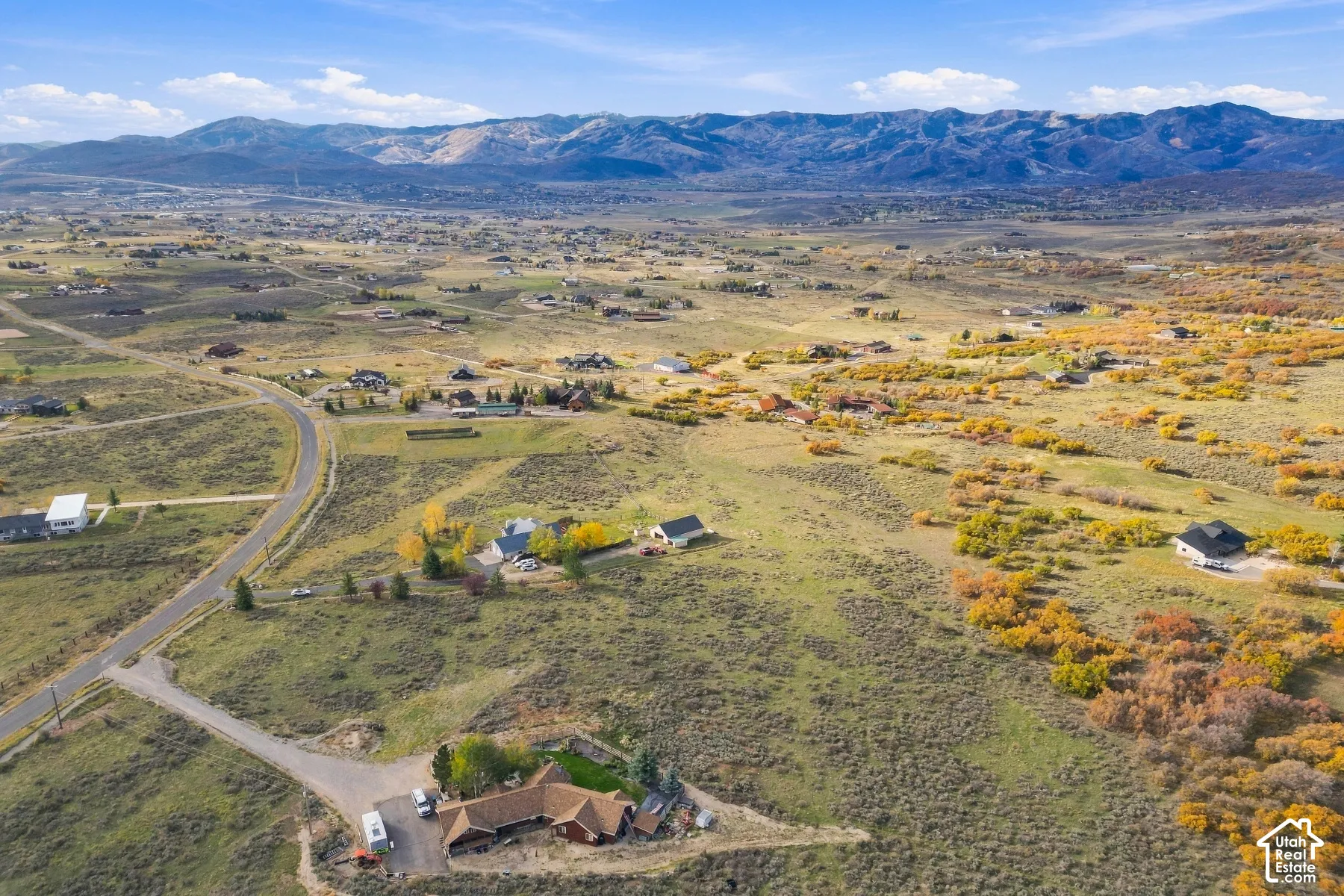 Aerial perspective of rural acreage featuring expansive terrain, paved driveway, mature trees, and mountain backdrop
