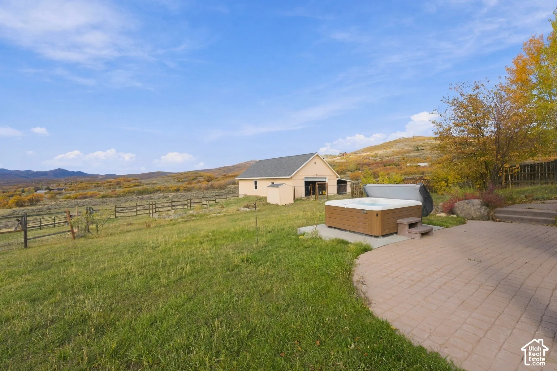 Expansive outdoor space featuring a paved patio, built-in hot tub, detached garage, and perimeter wood fencing, set against a mountainous backdrop