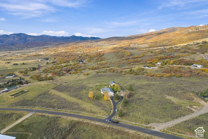 Aerial perspective of a multi-structure property featuring expansive acreage, paved asphalt driveway, mature deciduous trees, and surrounding mountain terrain