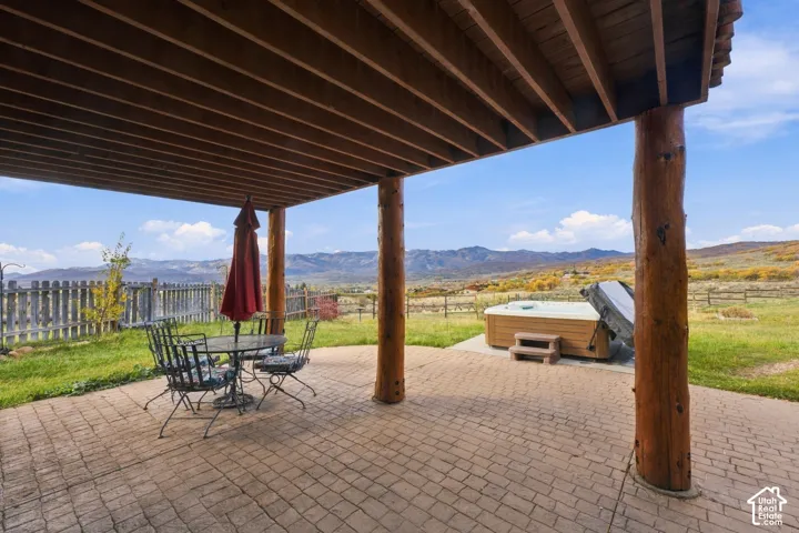Covered patio with rustic log columns and overhead timber beams, featuring stamped concrete hardscaping, an integrated spa, and mountain scenery