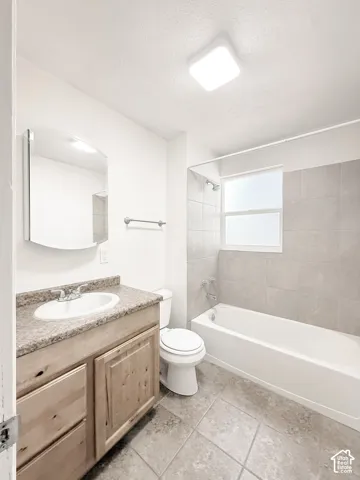 Bathroom featuring a natural wood-finish vanity, stone-patterned countertop with an integrated basin, tile flooring, and a tub-shower combination with floor-to-ceiling tile surround