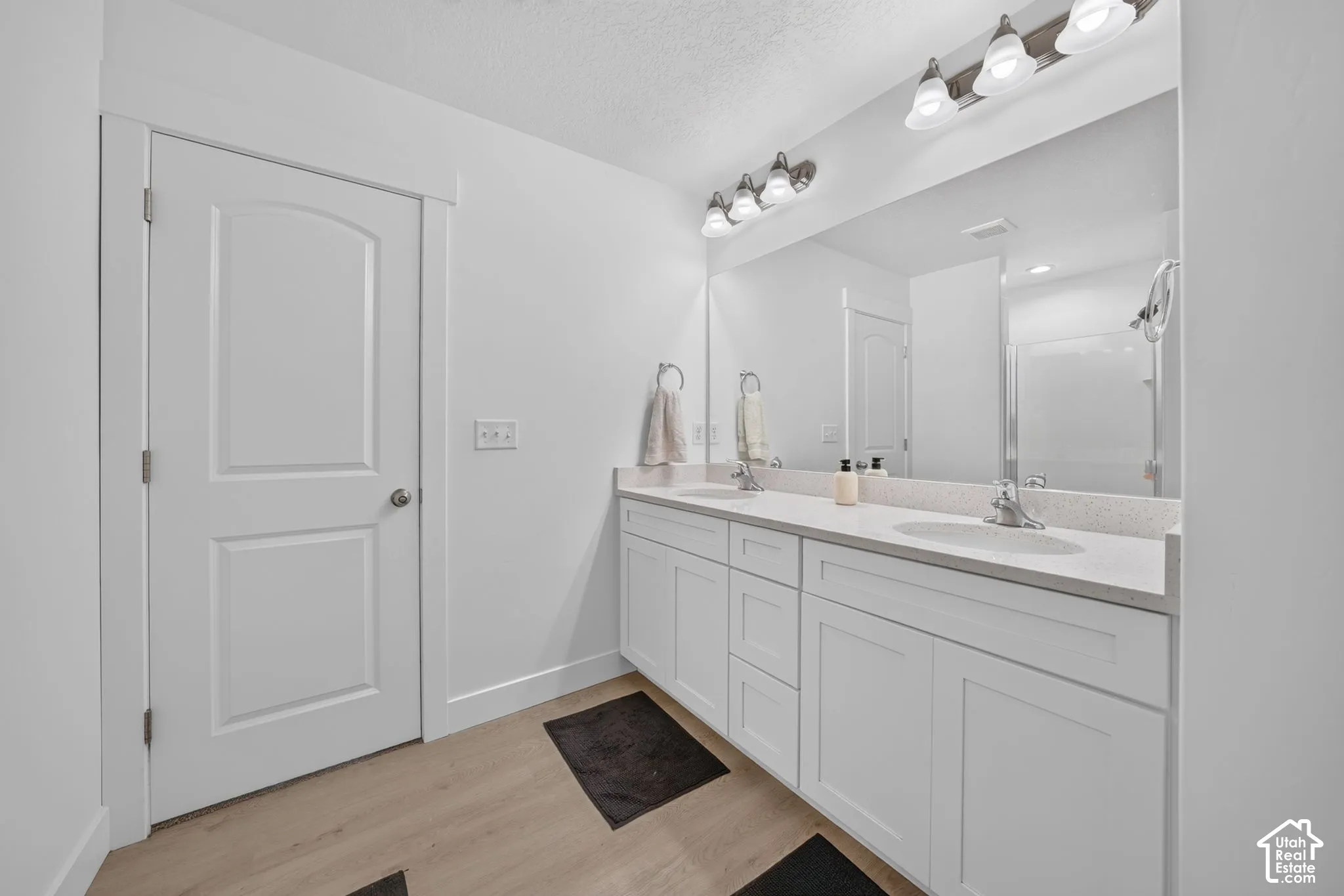 Dual-sink vanity with white shaker-style cabinetry, solid-surface countertop, full-width mirror, and two multi-bulb vanity light fixtures over wood-finish flooring