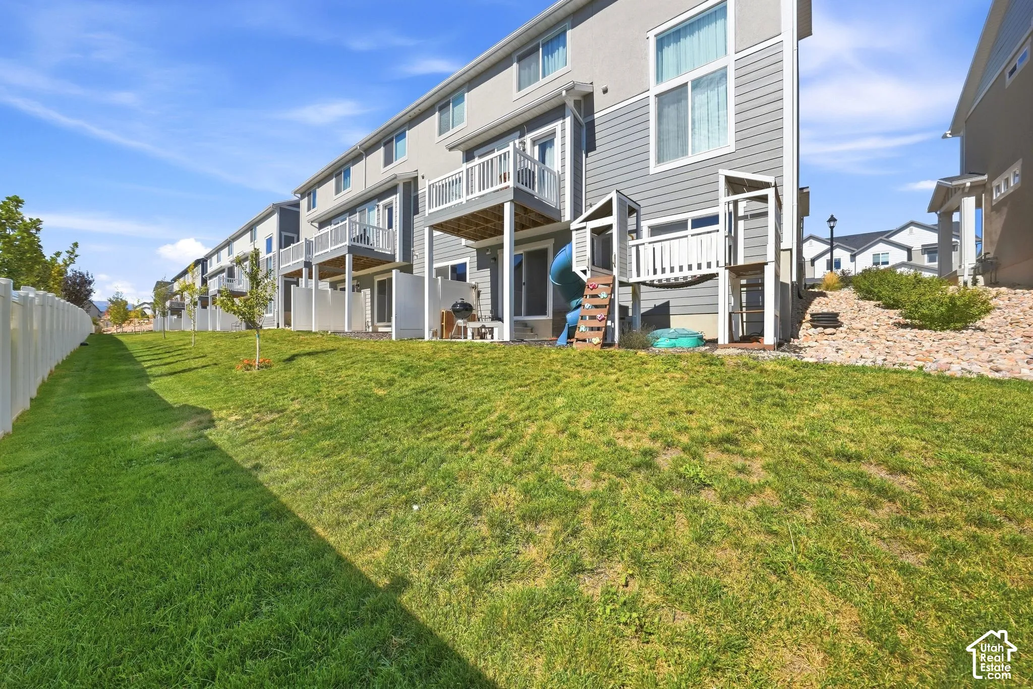 Multi-level residential exterior featuring horizontal siding, elevated balconies with white railings, and a sloped lawn with vinyl fencing