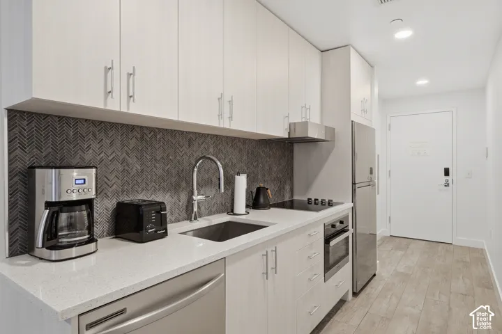 Kitchen featuring light-toned wood-finish cabinetry, stone-composite countertops, a herringbone-pattern tile backsplash, stainless steel appliances, and wood-finish flooring
