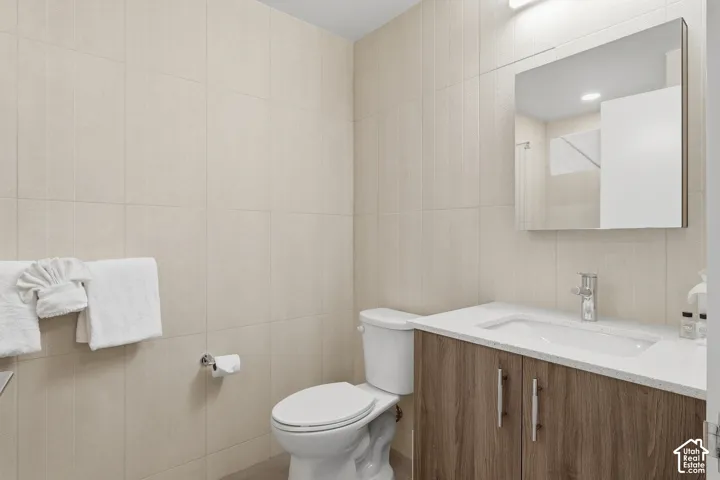 Bathroom featuring floor-to-ceiling neutral wall tile, vanity with wood-finish cabinetry, light-toned stone-composite countertop, rectangular undermount sink, and a frameless wall-mounted mirror