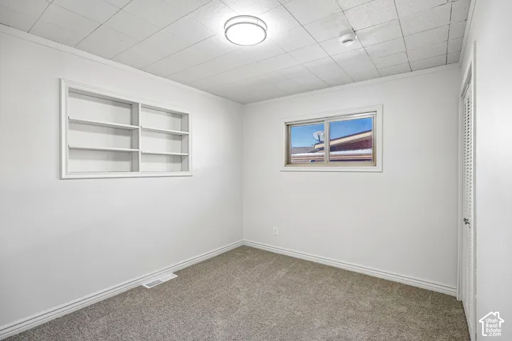 Room featuring neutral carpeting, a recessed wall shelving unit, a sliding window, a textured drop ceiling with flush-mount lighting, and a louvered closet door