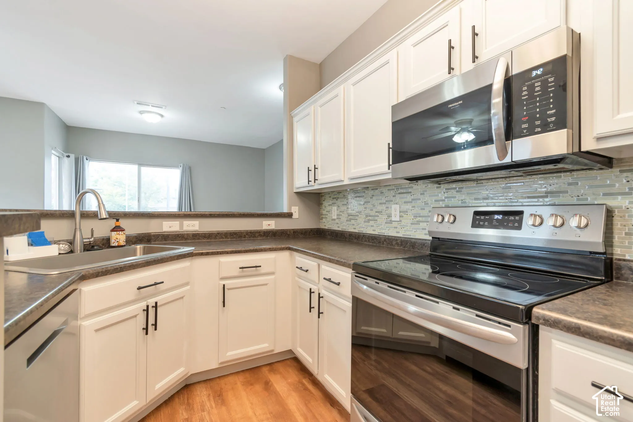 Kitchen featuring white shaker-style cabinetry, stainless steel appliances, a multi-tonal glass tile backsplash, dark laminate countertops, and wood-finish flooring