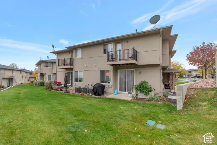 Rear exterior featuring stucco siding, two upper-level balconies with black metal railings, a concrete patio, and an expansive lawn