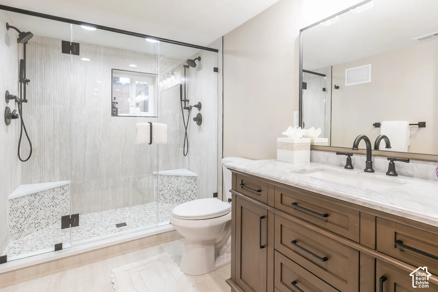 Walk-in shower featuring a glass enclosure, dual shower heads, and built-in tiled seating, complemented by a vanity with a stone countertop and dark metal hardware