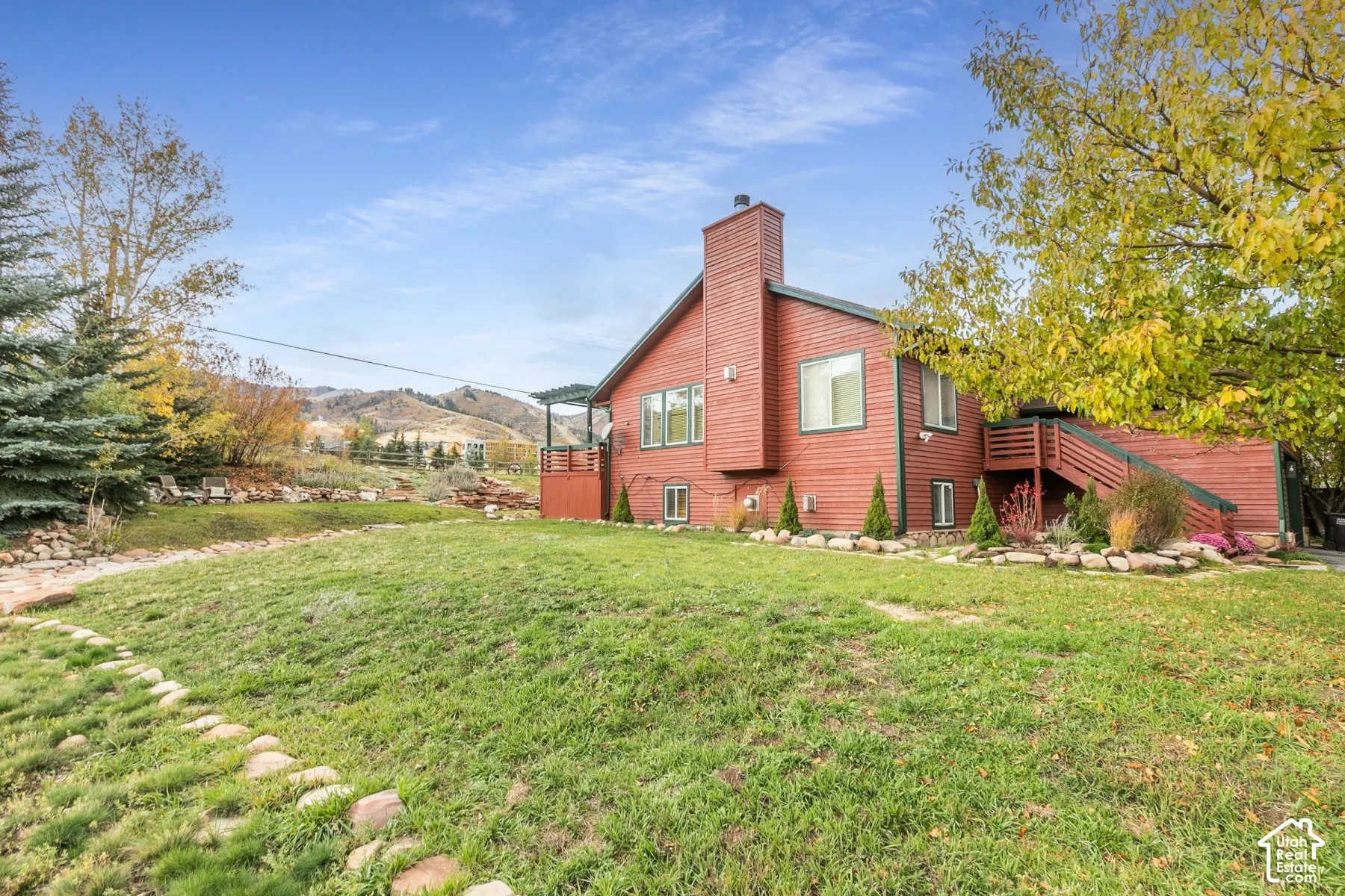 Exterior featuring wood-sided architecture, a prominent chimney, an elevated wooden deck, and a landscape with stone retaining walls and mature trees