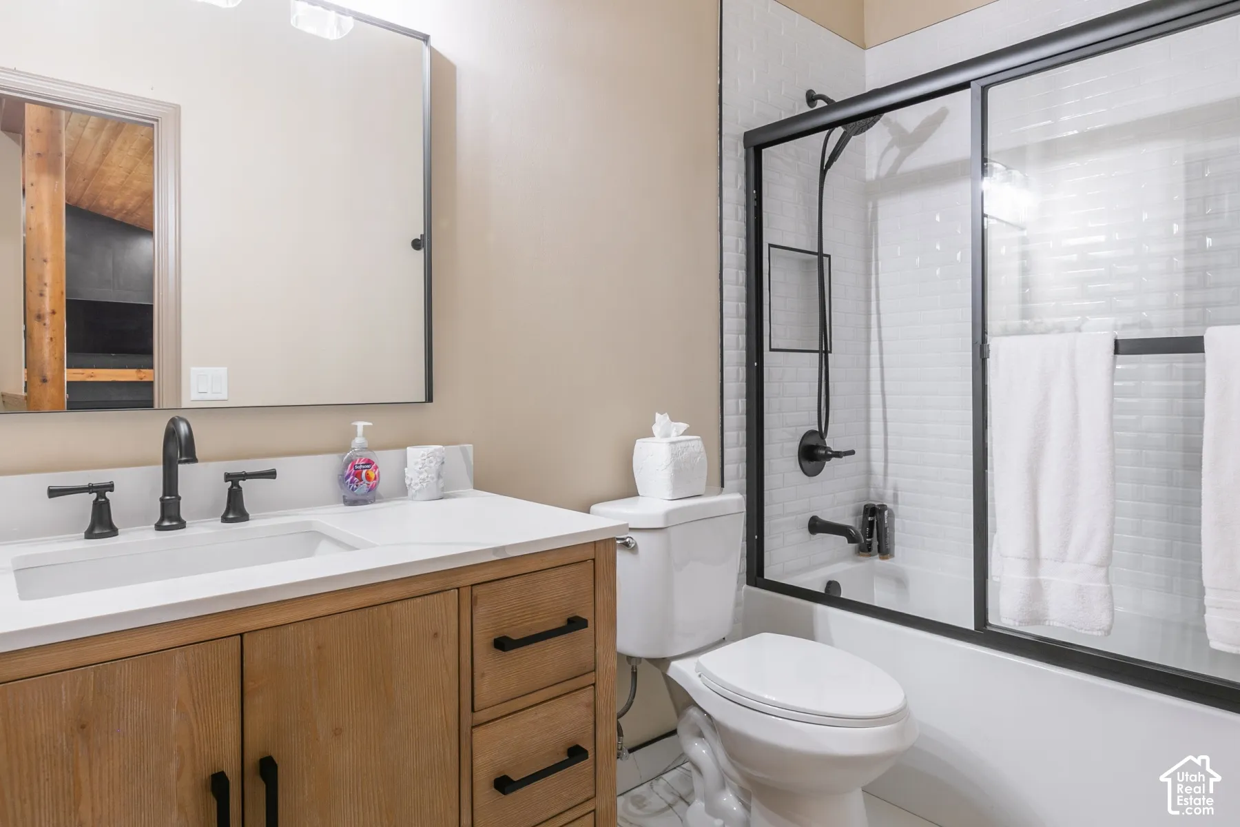 Bathroom featuring a wood-finish vanity with a white countertop, rectangular undermount sink, matte black hardware, and a tub-shower combination with subway-style wall tile and a framed glass enclosure
