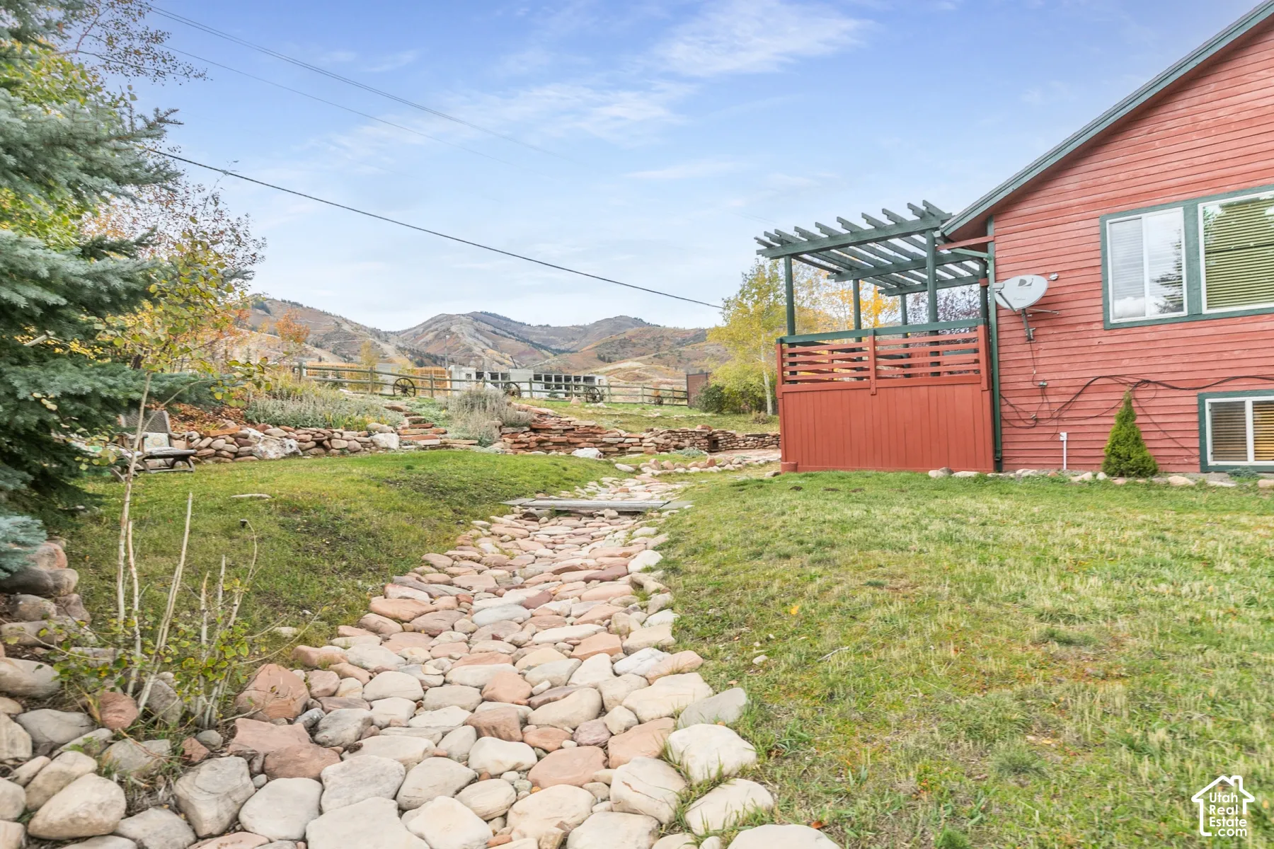 Exterior grounds featuring a decorative stone path, tiered natural stone retaining walls, an attached wooden pergola, and mountainous terrain in the background